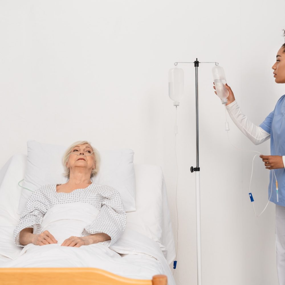 African american nurse holding bottle of intravenous therapy near senior patient in hospital